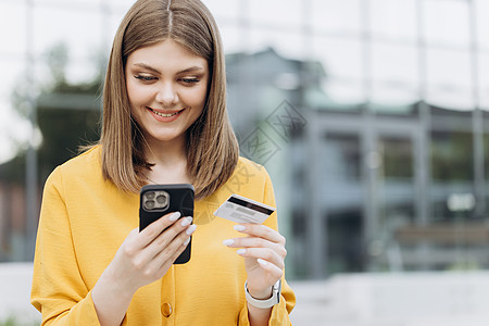 微笑的女孩拿着信用卡和智能手机坐在室外的长凳上 快乐的女性购物者使用即时便捷的移动支付在网上商店购物 电子银行应用程序服务背景图片