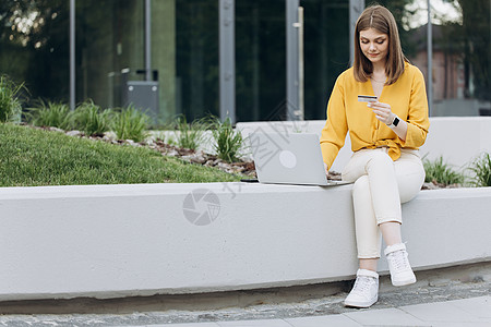 女性购物者使用即时便捷的移动支付在网上商店购物 电子银行应用程序服务 拿着信用卡和智能手机的女孩坐在室外的长凳城市建筑上背景图片