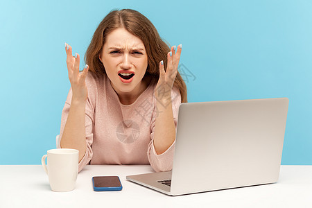 情感年轻女性在蓝背景的笔记本电脑上工作商务女士人士商业博主自由职业者愤怒手势尖叫老板图片