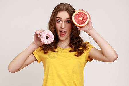 穿着黄色T恤的嬉戏性兴奋少女拿着一半成熟多汁的葡萄油和圆甜甜圈 用张开嘴看相机图片