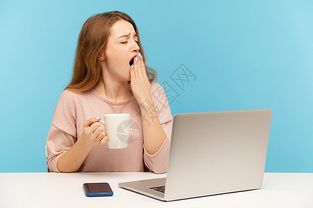 情感年轻女性在蓝背景的笔记本电脑上工作活力员工女士杯子办公室失眠时间咖啡唤醒就寝图片