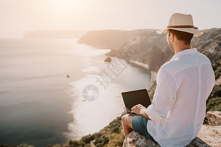 数字游牧者 自由职业者 假期远程工作 戴帽子的男人 一个带着笔记本电脑的商务人士 在日落时分坐在海边的岩石上 从远处在网上进行商图片