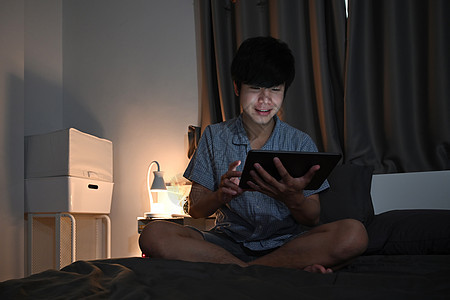 微笑的男人检查社交媒体 在数字平板电脑上 卧室的夜间图片