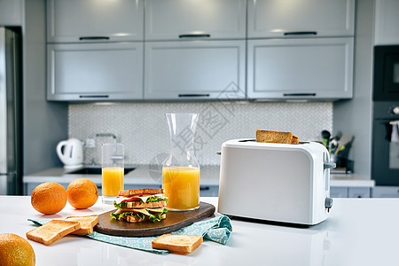 餐桌上有橙汁和三明治的白色电动面包面包机 在舒适的厨房里吃早餐橙子营养工具窗户甜点器具果汁炙烤烤面包机水果背景图片
