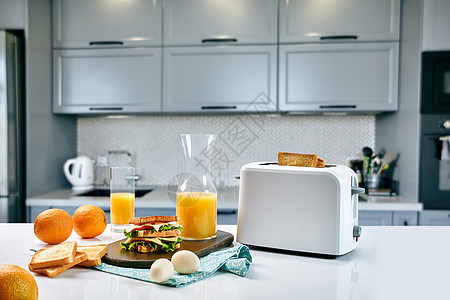 餐桌上有橙汁和三明治的白色电动面包面包机 在舒适的厨房里吃早餐咖啡桌子机器玻璃炙烤橙子果汁水果烤面包机窗户背景图片