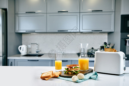 餐桌上有橙汁和三明治的白色电动面包面包机 在舒适的厨房里吃早餐电气橙子机器花朵水果炙烤工具木头器具制作者图片