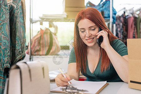 年轻的女商务人士面带微笑 在她的服装店办公室用智能手机与顾客交谈 并记下订单 工作和经营理念图片