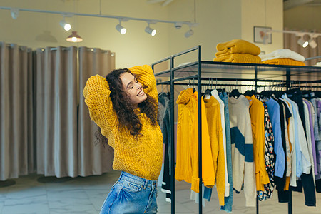 一位年轻漂亮的卷发女人的画像 穿着黄色保暖毛衣和牛仔裙在一家服装店里摆姿势 穿着和摆姿势 背景是衣架上的配饰 展示 吹牛 看着镜背景