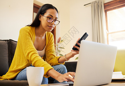 在明亮的客厅里 一位女士坐在沙发上远程工作 一边在笔记本电脑上打字 一边拿着智能手机 一位专注于使用现代技术在家戴眼镜的西班牙裔图片