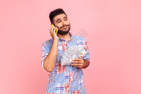 有魅力的年轻成年男子的画像 留着胡子 身穿蓝色衬衫 站着 拿着钱扇 在手机上聊天 吹嘘利润图片