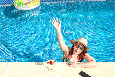 一个红头发的年轻女人微笑着 从游泳池里挥手 在阳光明媚的日子里冷静下来 暑假在泳池边晒日光浴的年轻女孩 夏季和休闲时间的概念图片