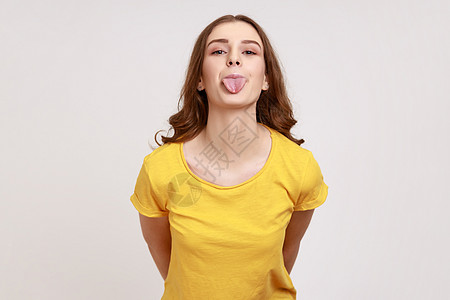 穿着黄色T恤的年轻女性 带着幼稚面部表情和舌头露出笑柄 看着镜头 看相片图片