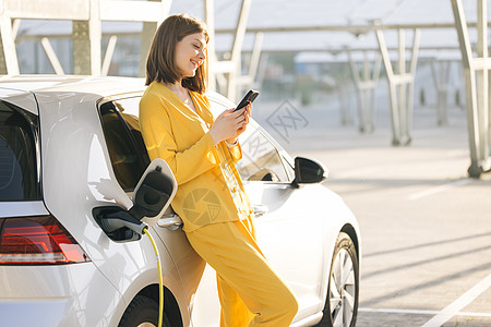 在太阳能发电厂附近的充电站为电动汽车充电的白人女性在等待时使用智能手机图片