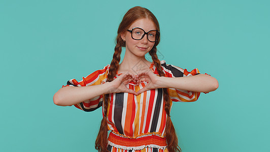 微笑的姜女做心脏手势展示爱的标志表达好感和同情孩子机构背景享受蓝色女性喜悦浪漫红发广告背景图片