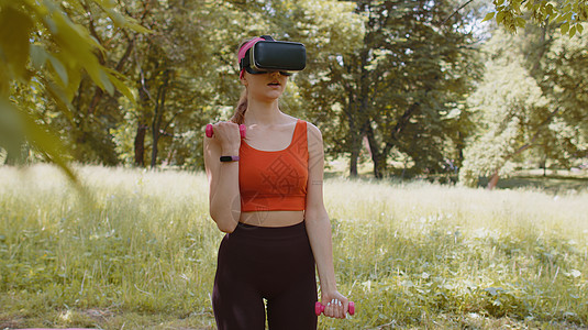 VR头盔耳头盔的听风女孩 在公园户外用哑铃进行健身锻炼运动背景图片
