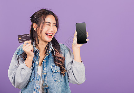 妇女自信地穿戴尼姆牛仔裤衬衫 微笑地展示手机空白屏幕和信用卡银行电话女性购物细胞借方支付女士工作室信用紫色图片