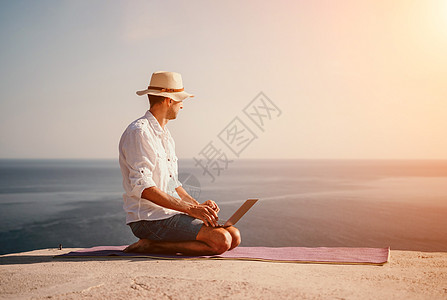 数字游牧者 自由职业者 假期远程工作 戴帽子的男人 一个带着笔记本电脑的商务人士 在日落时分坐在海边的岩石上 从远处在网上进行商图片