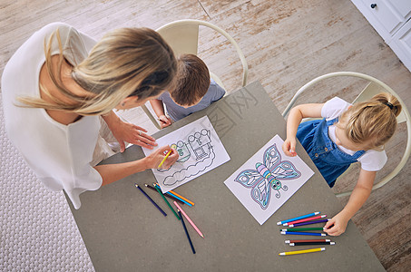 上图是小女孩和男孩坐在桌旁 拿着彩色铅笔和图片 妈妈在帮忙涂色 有两个孩子的白人母亲享受教育消遣和创造力图片