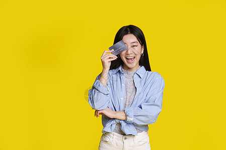 兴奋的亚洲女孩微笑着用银行卡右眼遮住她准备花钱 身穿蓝色衬衫的亚洲女孩 银行卡样机在黄色背景下被隔离 购物理念图片