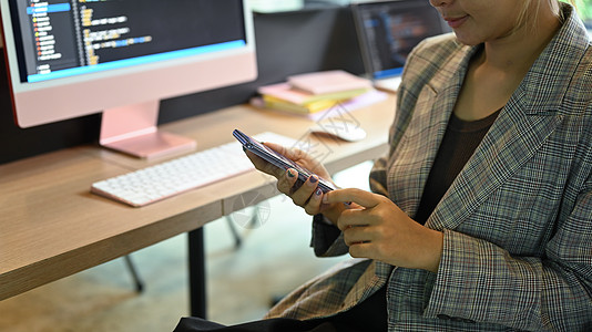 使用手机的女性软件开发人员 坐在办公桌上 电脑屏幕上有编程代码图片