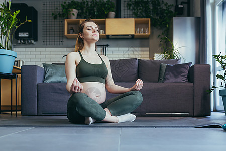 怀孕的美孕妇在家做瑜伽 坐在莲花姿势的运动垫子上健身房运动装腹部平衡训练母亲身体眼睛练习药品图片