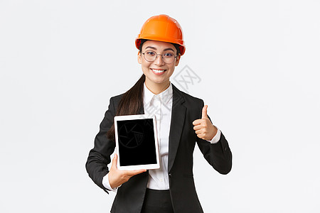 专业持微笑的像亚洲女性建筑经理 身穿商业服和戴安全头盔的工厂工程师 用正面图表 白背景图显示拇指上方和数字平板屏幕建设者维修办公图片