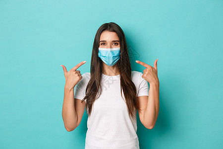 流行病 covid-19 和社会距离的概念 身穿白色 T 恤 微笑友好的女孩 建议在冠状病毒期间戴上医用面具 站在蓝色背景上图片