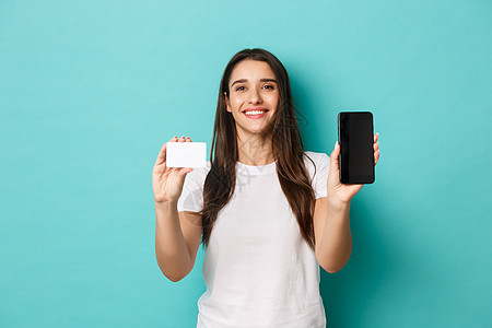 快乐迷人的女模特肖像 展示手机屏幕和信用卡 微笑着高兴 站在蓝色背景下图片