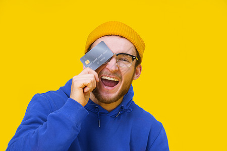 带着信用卡 手持借记卡微笑着遮住他的眼睛 准备花钱 身穿蓝连帽衫的年轻人拿着假冒银行卡 在黄色背景中被孤立 购物概念图片
