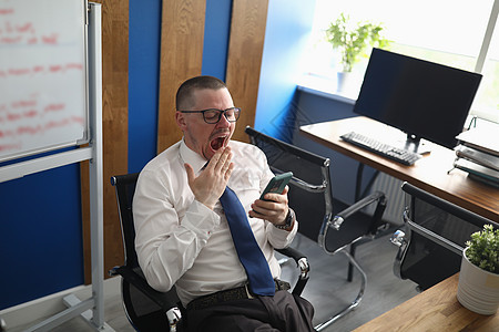 商务人士在工作场所寻找智能手机和打哈欠机成人商业眼镜老板男人工作失眠办公室首席职员图片