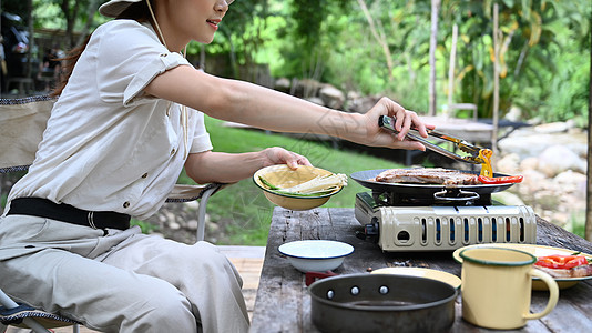 女性旅行者在河岸附近露营时坐在折叠椅和烤烤肉边坐着 户外活动或休假概念图片