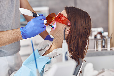 专业男医生在现代牙科柜中为快乐的白人女性补牙后使用紫外线灯治疗眼镜病人药品牙医程序激光化妆品矫正口服图片