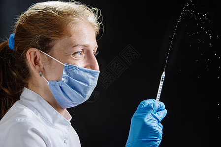 医生在黑色背景下用注射器喷药 医生在他的防护面具下微笑 注射器中的药物倾泻到空气中 一位戴着医用口罩 手套和蓝色背景大注射器喷液图片