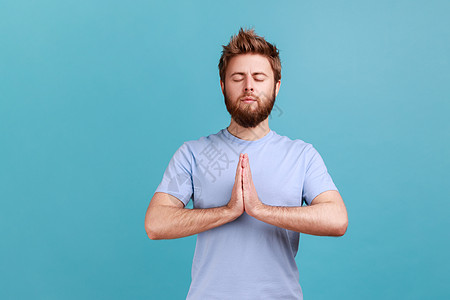 穿着蓝色T恤的男子站在闭着眼睛和祈祷手势 冥想 集中思想的面前图片