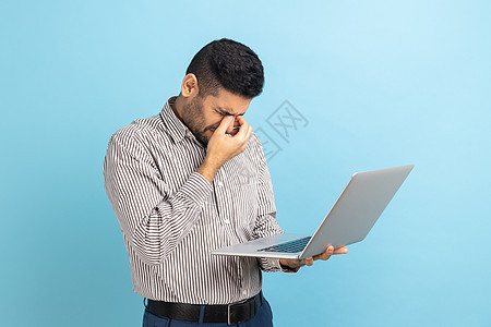 商务人士拿着笔记本电脑站在手边 摩擦他的眼睛 看起来很疲惫和疲劳图片