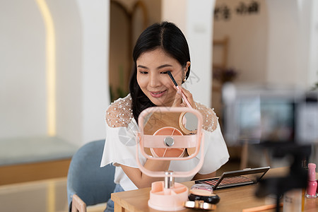 亚洲女性美容博主直播在社交媒体上评论化妆品 现代年轻女性影响者在智能手机摄像头上展示她的日常化妆品魅力互联网居住刷子记录技术营销图片