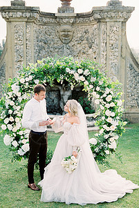 Groom在花园的婚礼拱门上 穿着白色礼服 把戒指戴在新娘的手指上图片