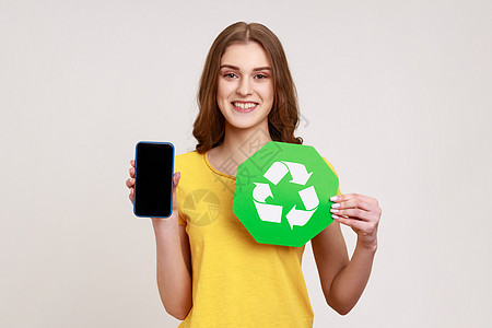 身穿黄色 T 恤的快乐迷人少女拿着带空白屏幕和回收符号环境图标的手机 看着相机微笑图片