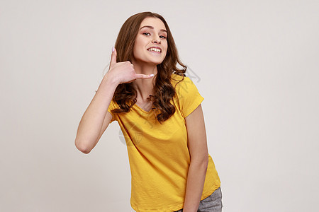 打给我 穿黄色短袖T恤的快乐嬉戏少女的肖像 在电话手势上说话 微笑 调情 问话图片