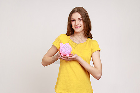 带着棕色头发的迷人笑脸少女拿着小猪银行 乐于积蓄钱 穿着黄色休闲式T恤衫图片