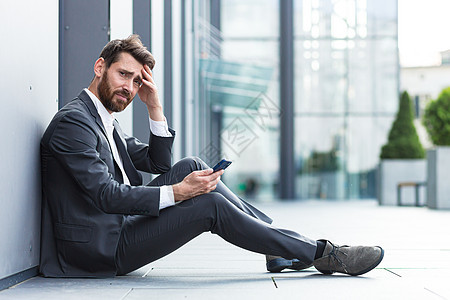 坐在户外办公室附近的办公室职员得到坏消息 沮丧和疲倦读坏消息男性职业电话商务手机危机地面阅读压力商业图片