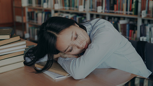 年轻美丽疲惫的亚洲女学生坐在书桌前 头靠在书桌上 睡在大大学图书馆里被一堆堆东西包围的教科书上图片