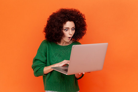 身穿绿色散装毛衣的非洲发型女性在便携式电脑上工作 透过张开嘴看露台 她被震惊地描绘成一幅肖像图片