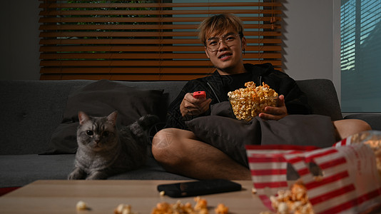 快乐的年青年轻男人在看电视吃爆米花时 与可爱的猫坐在家舒适的沙发上图片