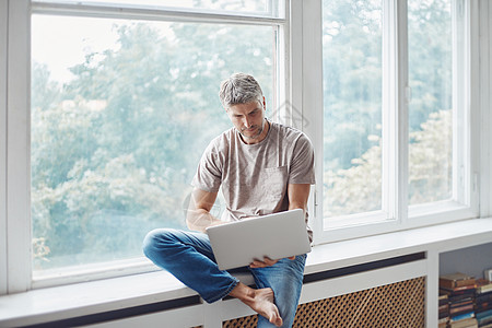 男人用笔记本电脑坐在起居室的窗台上图片