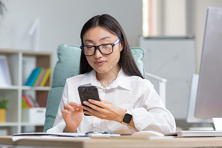 一位戴眼镜的亚洲女商务人士手里拿着电话 拨打一个信息 笑声女士公司管理人员电子邮件服务微笑办公室职业商务阅读图片