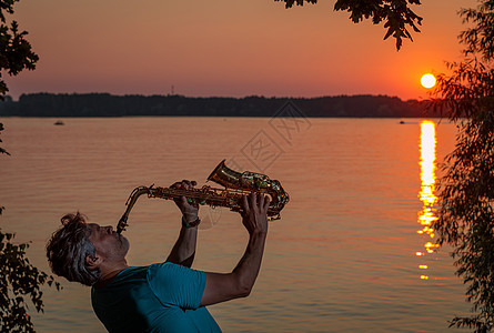 一个成年男子在日落时 晚上在河边玩萨克斯金子木管男性阴影音乐家旋律太阳反射黄铜艺术家图片