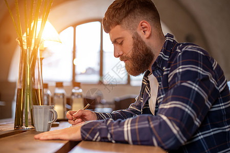 年青的留胡子商务人士坐在咖啡馆里 坐在桌子上用笔记本写作潮人阅读成人电子书人士办公室日记药片学生商务图片