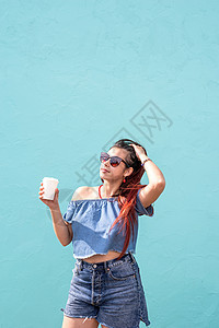 在街上蓝墙背景的红发 喝着咖啡跳舞的时尚女性在街头欢喜太阳镜头发喜悦成人蓝色快乐饮料电话女孩衣服图片
