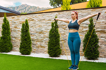 年轻女运动员将身体肌肉伸展到户外她家花园里的弹性带 关于健康和福祉的概念 请查看InfoFinland上的“健康与幸福”图片